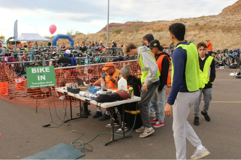 Race volunteers in high-visibility vests at a table near a bike transition area with a 'BIKE IN' sign.