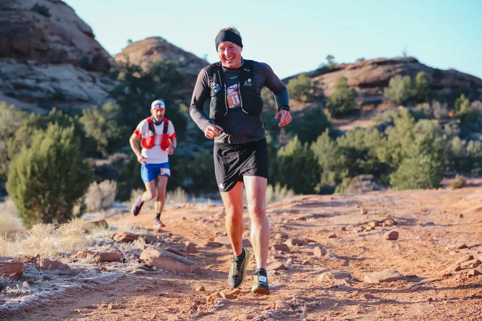 A smiling runner in the foreground runs on a dusty trail with another runner behind him, with red rock formations and green bushes in the background.