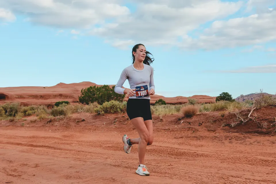 A female runner with bib number 1105 runs on a dusty red trail with desert scenery and a blue sky with clouds in the background.