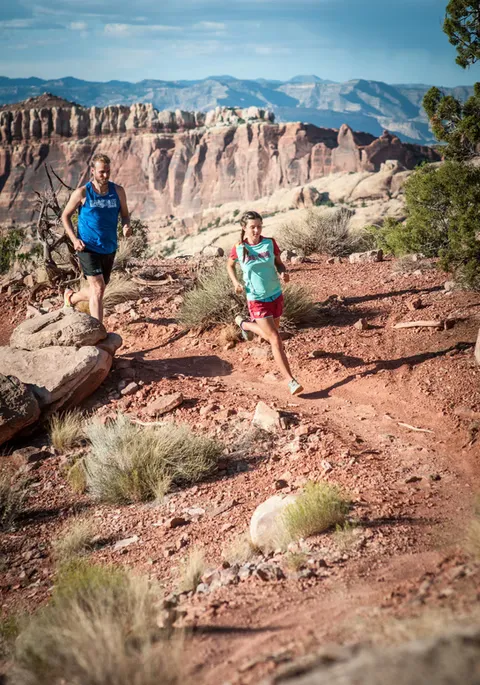 Two runners, a man and a woman, race down a rocky, desert trail with a dramatic canyon landscape in the background.