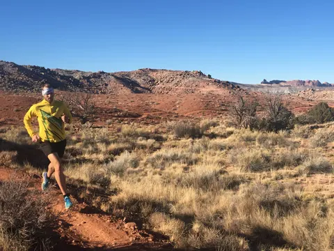 A runner in a yellow shirt and black shorts runs on a dirt trail with red rock formations and desert vegetation in the background under a clear blue sky.