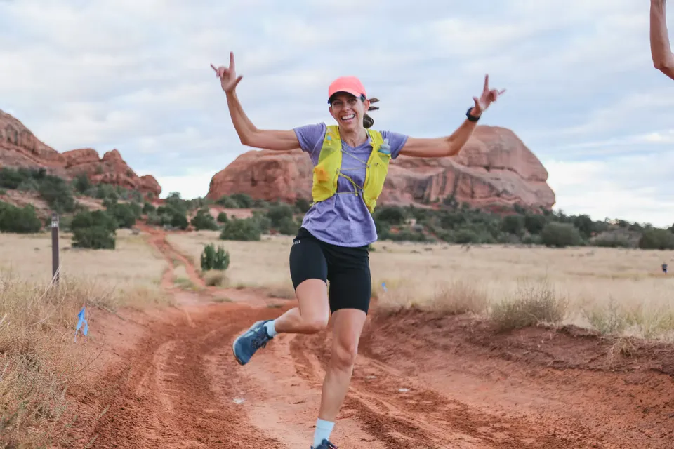 A female runner with arms raised in celebration runs on a red dirt trail with scenic rock formations in the background.
