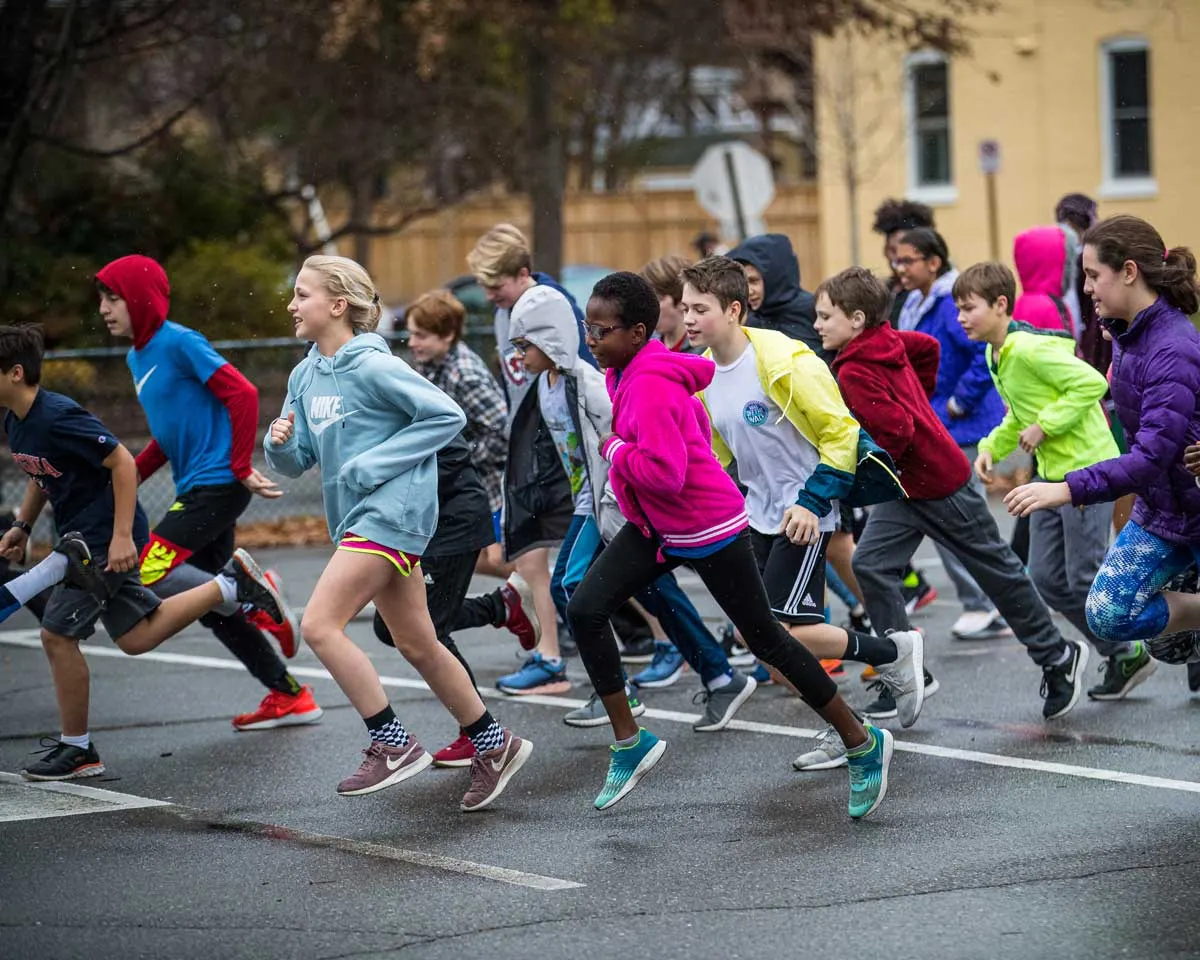 A group of children running in a race, with some wearing athletic clothing and others in hooded jackets.