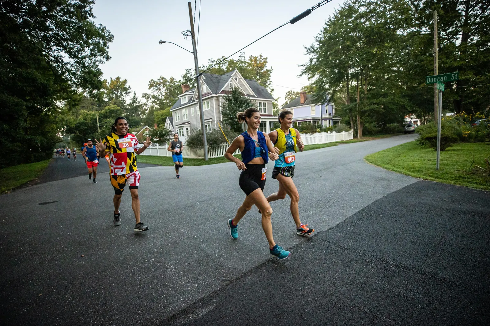 Runners participating in a race on a paved road with houses and trees in the background.