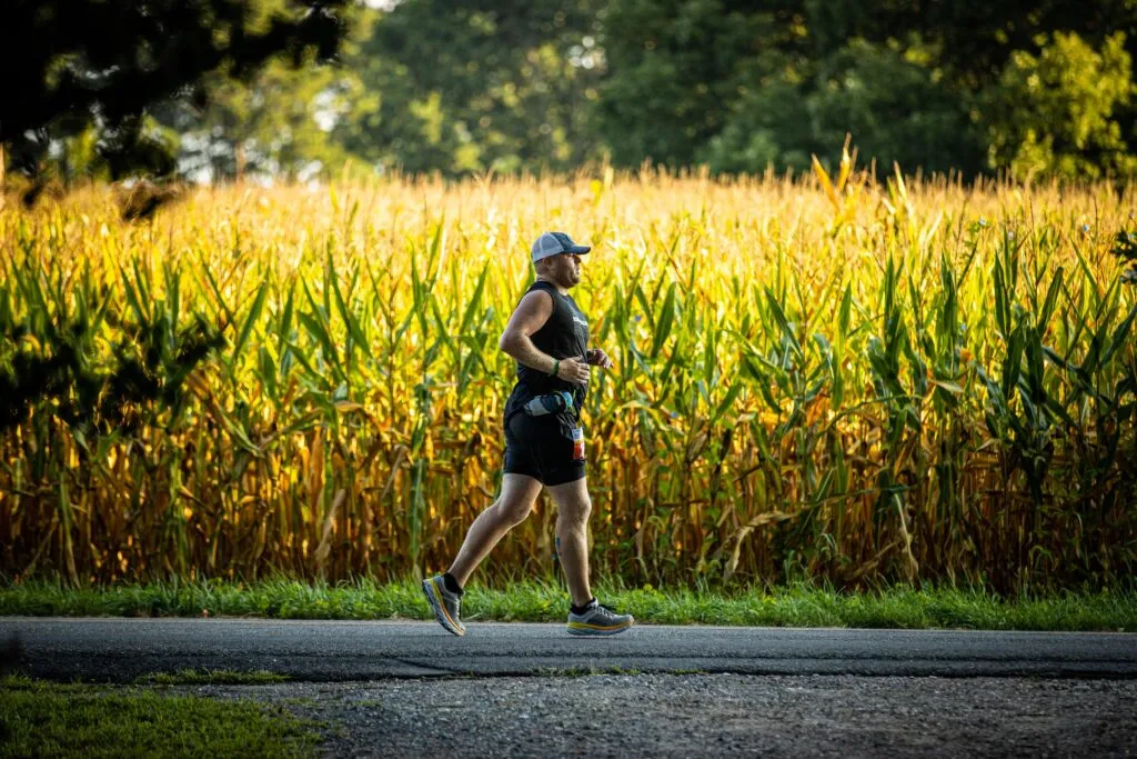 A male runner runs on a paved road with a large cornfield to his right and trees in the background.