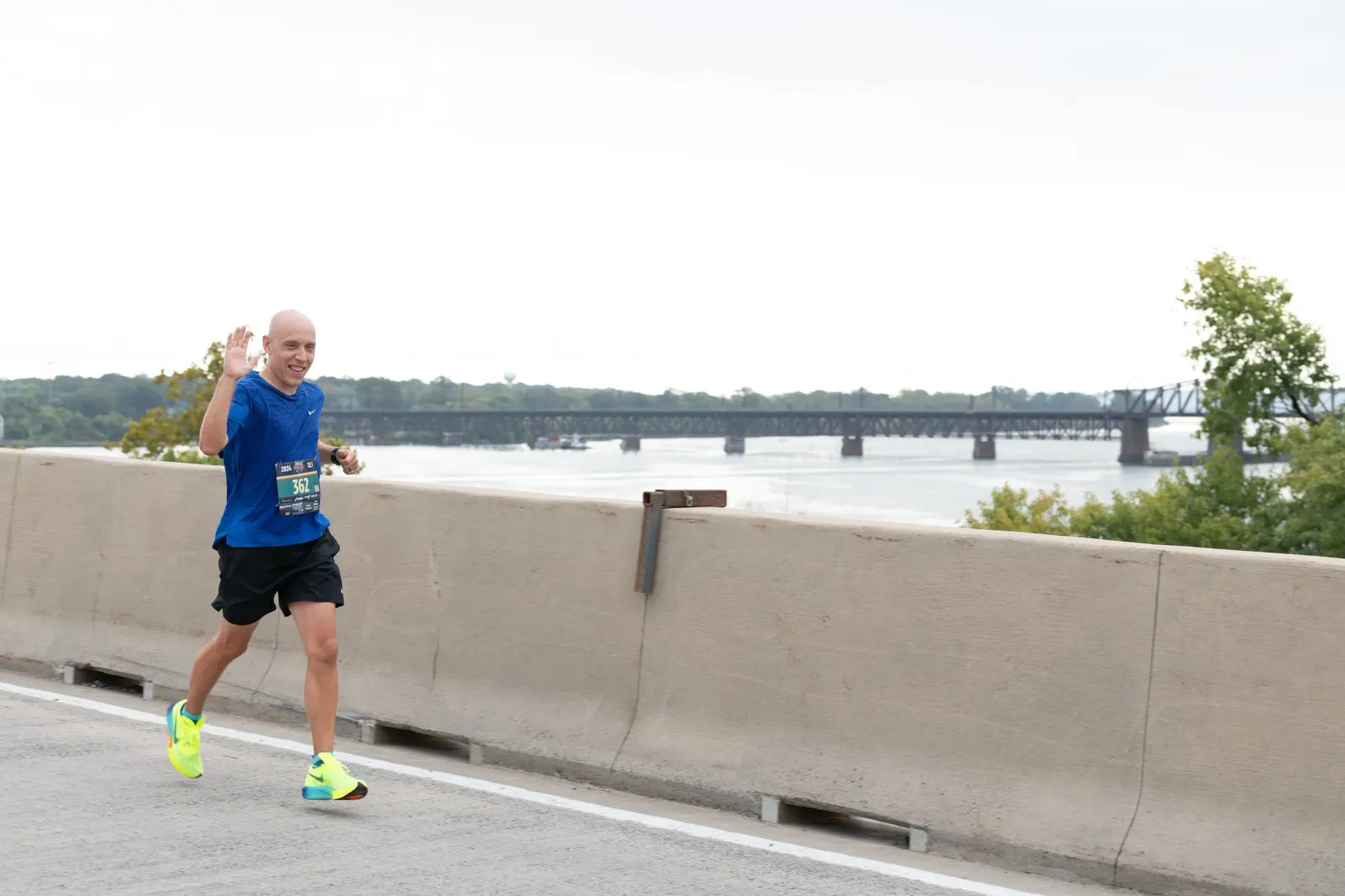 A male runner with a shaved head, wearing a blue t-shirt and black shorts, waves as he runs on a road with a concrete barrier. A bridge and a body of water are visible in the background.