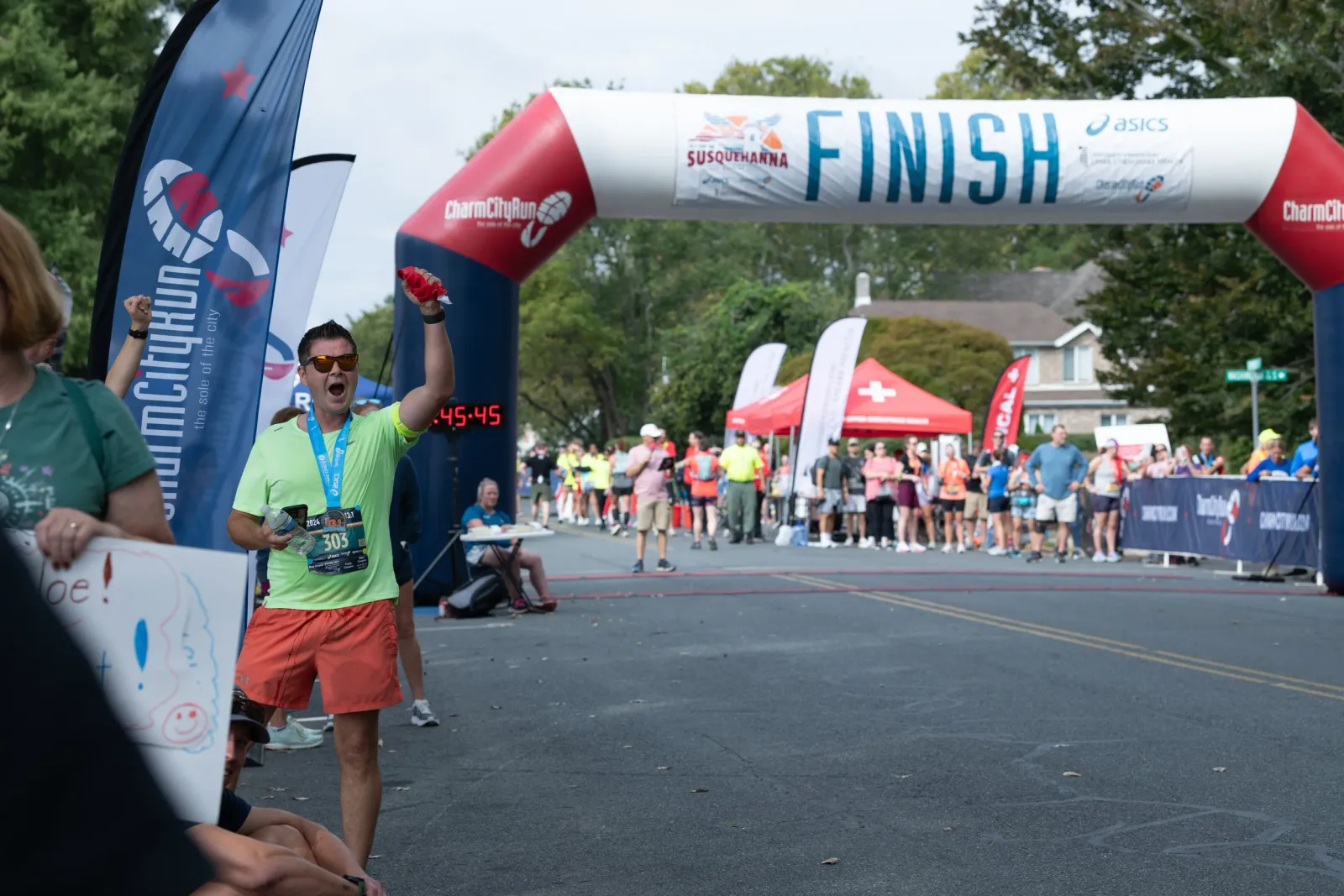 A runner in a neon green shirt and orange shorts celebrates as he crosses the finish line, raising a fist in the air. A large inflatable finish line arch is visible behind him, with other runners and spectators in the background.