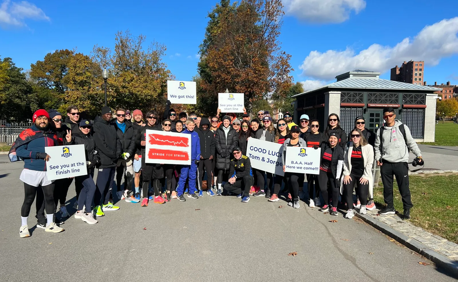 A group of runners posing for a photo, holding signs that say 'We got this!', 'See you at the start line', 'Stride for Stride', 'GOOD LUCK Tom & Jorge', and 'B.A.A. Half Here we come!!!'.