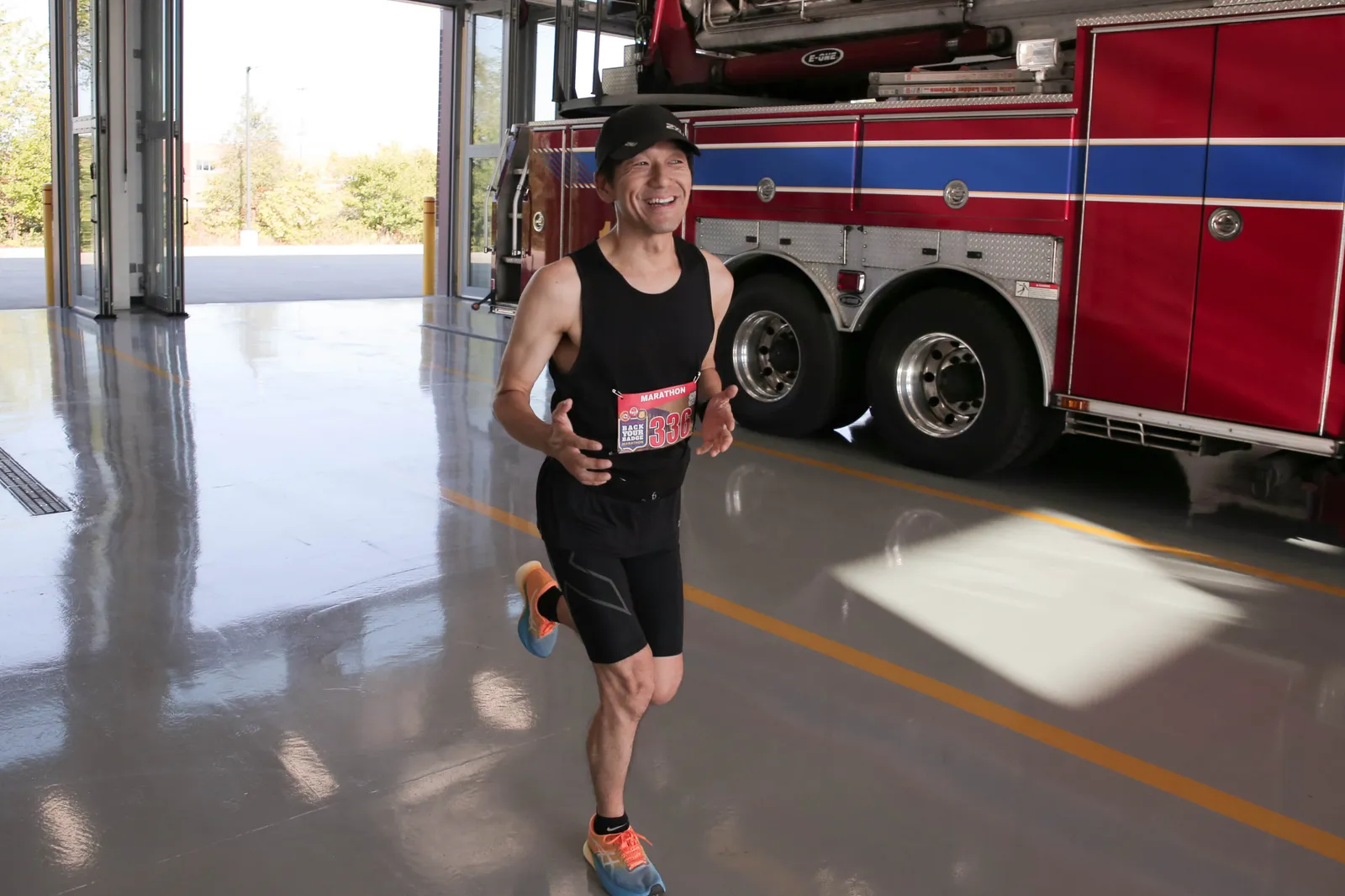 A smiling male runner wearing a black tank top, black shorts, and a race bib numbered 336, running in front of a red fire truck.