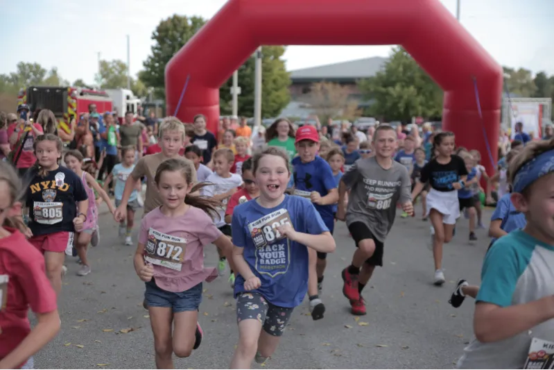 Children running towards the camera under a red inflatable arch at the start of a kids' fun run.