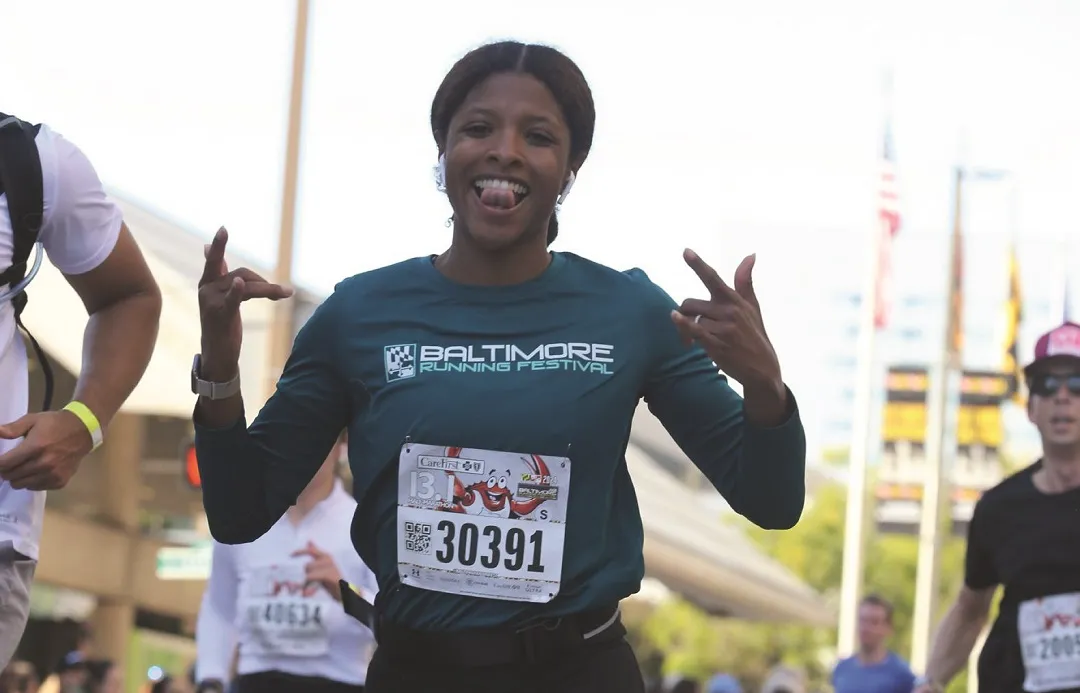 A female runner with a big smile and tongue sticking out, wearing a Baltimore Running Festival shirt and bib number 30391, makes a celebratory gesture while running.