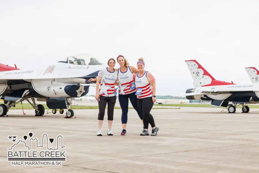 Three women in race attire pose in front of United States Air Force fighter jets at the Battle Creek Half Marathon & 5K.