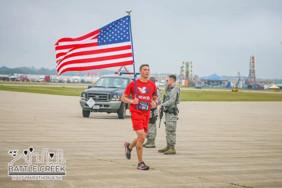 A runner carries an American flag during the Battle Creek Half Marathon & 5K, with military personnel and carnival rides in the background.