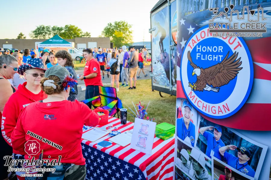 People gathered at the Battle Creek Half Marathon & 5K event, with volunteers at a table and race branding visible.