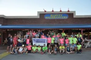 A large group of runners in athletic wear pose for a photo in front of a building with 'HARVEY WASHBANGER's' signage.