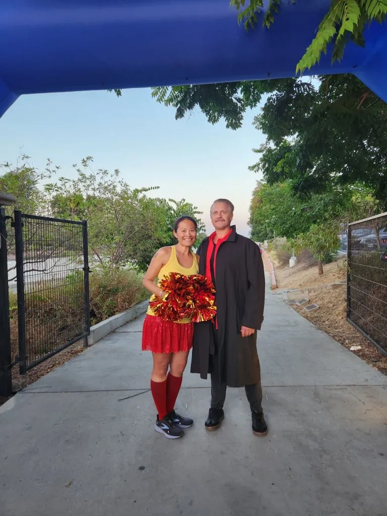 A man and a woman in costumes, one holding pom-poms, standing under a blue archway at a race event.