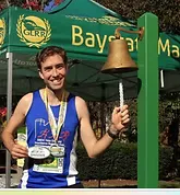 A smiling runner wearing a race bib and multiple medals rings a bell under a tent that says Baystate Marathon.