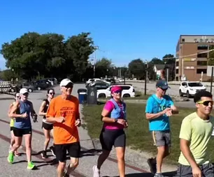 A group of runners of various ages and genders run along a paved path with trees and buildings in the background.