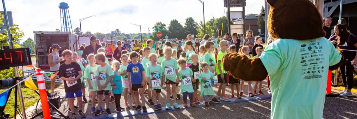 A group of young runners lined up at the start of a race, with a person in a bear mascot costume facing them.