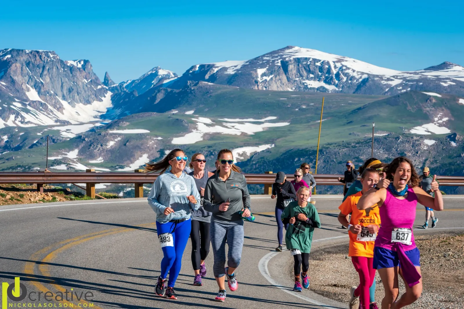 Runners race along a mountain road with snow-capped peaks in the background.