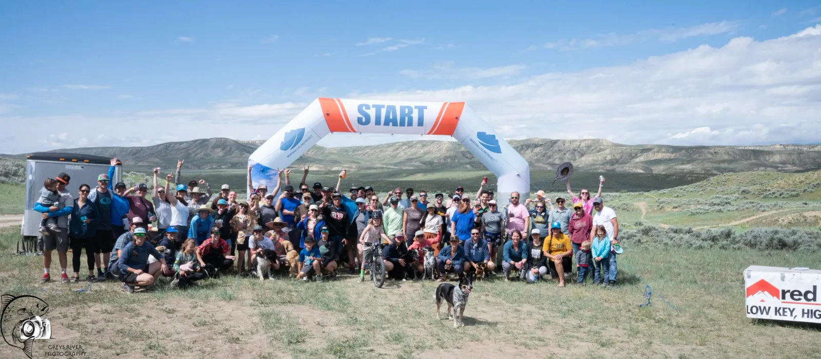 A large group of runners and spectators pose for a photo under a start line arch, with rolling hills in the background.