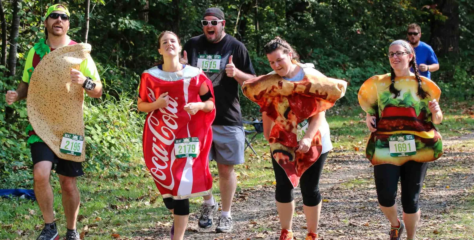 Runners in food-themed costumes, including a taco, Coca-Cola can, pizza slice, and hamburger, participating in a race.