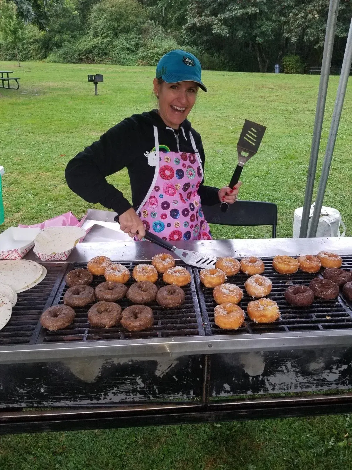 A woman wearing a blue baseball cap and a pink donut-themed apron smiles while grilling donuts outdoors.