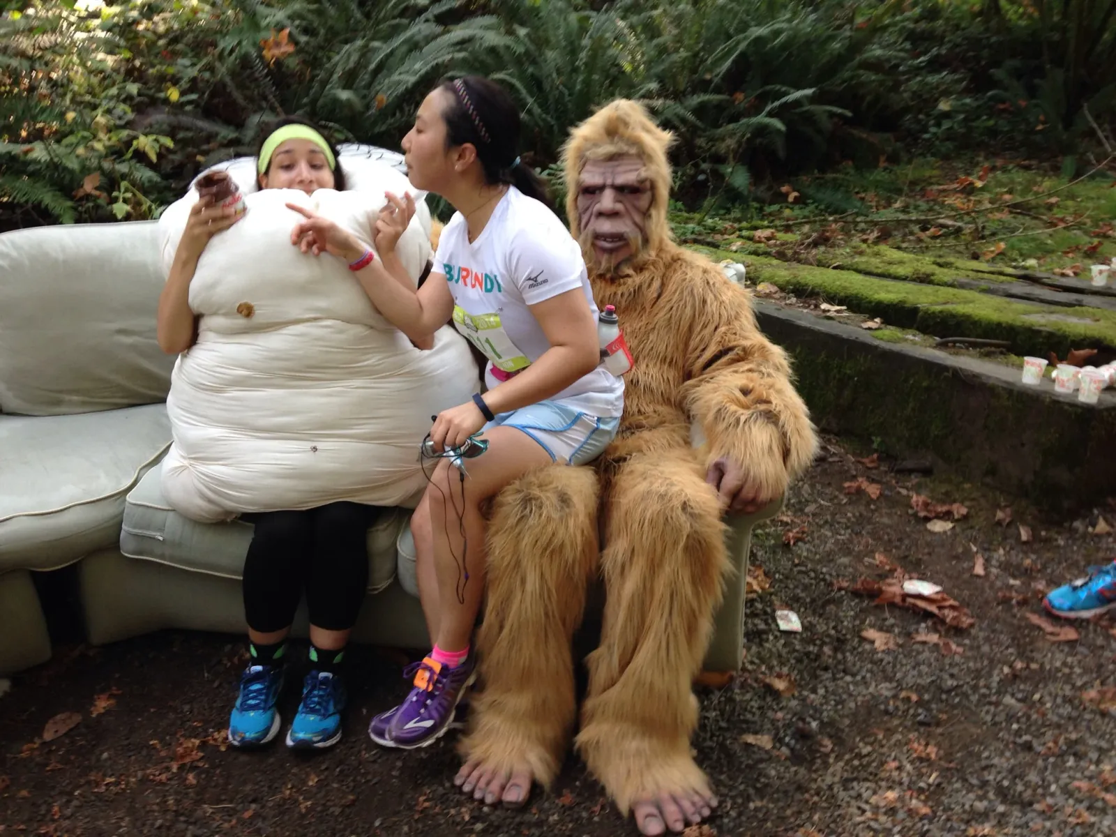 A person in a Sasquatch costume sits on a couch next to another person dressed as a giant marshmallow, both appearing to be at a race event.