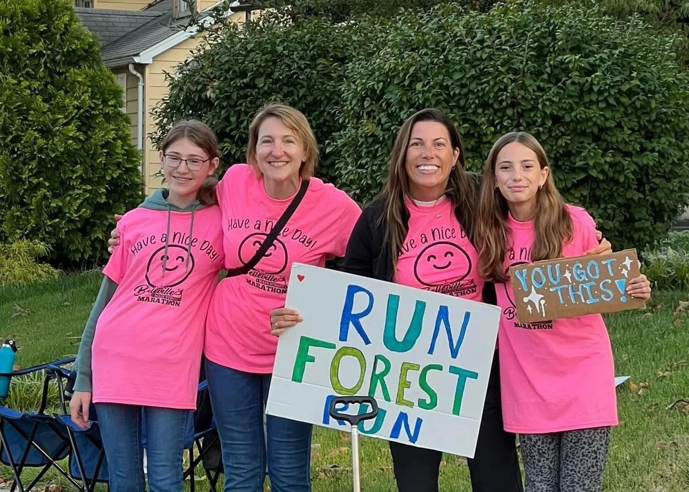 A group of four people, two adults and two young females, wearing pink t-shirts with race logos and smiling. They are holding signs that say "RUN FOREST RUN" and "YOU GOT THIS!"