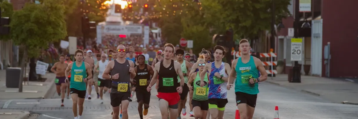 A group of runners are racing down a street lined with trees and buildings, with a finish line visible in the distance.