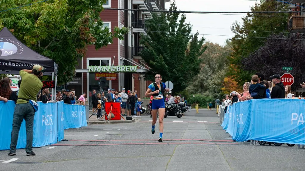 A female runner crosses the finish line of the Bellingham Bay Marathon, with spectators and buildings in the background.