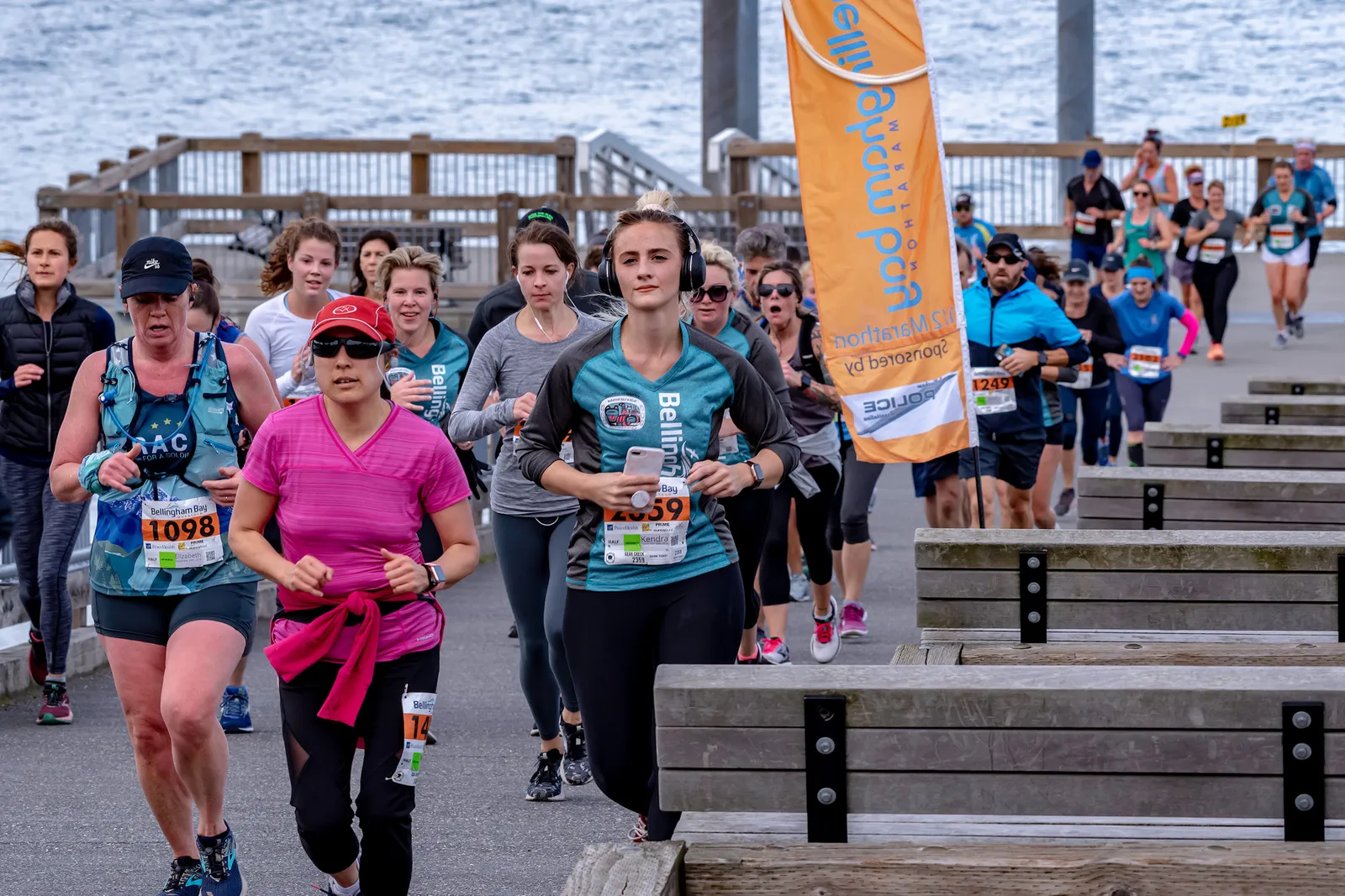 A group of female runners in the foreground and background, some wearing headphones, running along a paved path next to a wooden boardwalk. An orange banner with text is visible on the right side of the image.