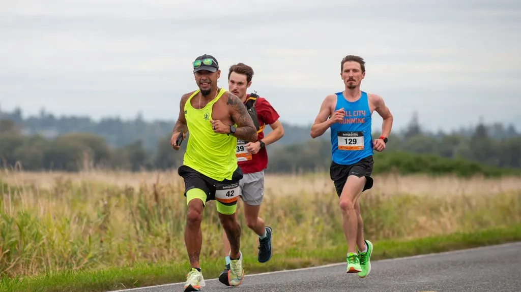 Three runners race along a road, with the lead runner in a bright yellow tank top, the second in a red shirt, and the third in a blue tank top.