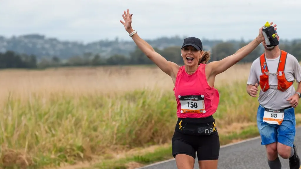 A female runner with her arms raised in celebration, wearing a pink tank top and black shorts, running a race with a male runner behind her.