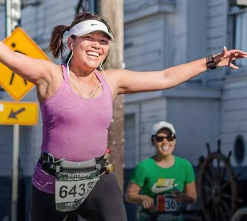 A smiling female runner in a purple tank top and black leggings runs with her arms outstretched, wearing a race bib with the number 6433.