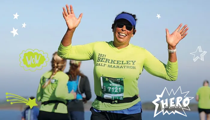 A female runner wearing a bright green Berkeley Half Marathon shirt smiles and waves with both hands raised as she runs, with other runners in the background.