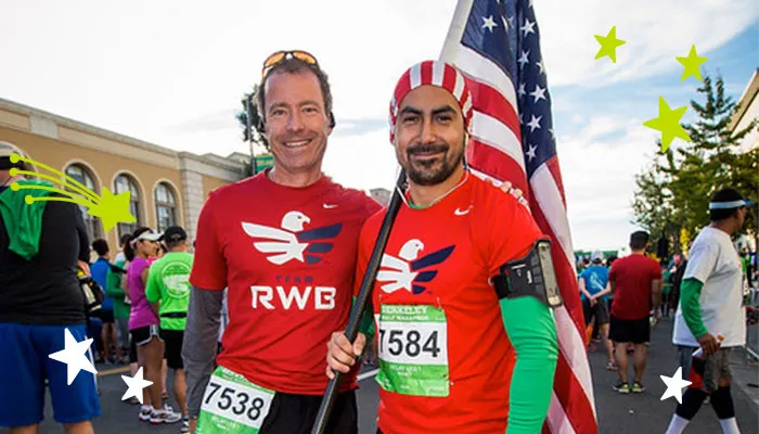 Two runners smiling, one holding an American flag, at the start of a race.
