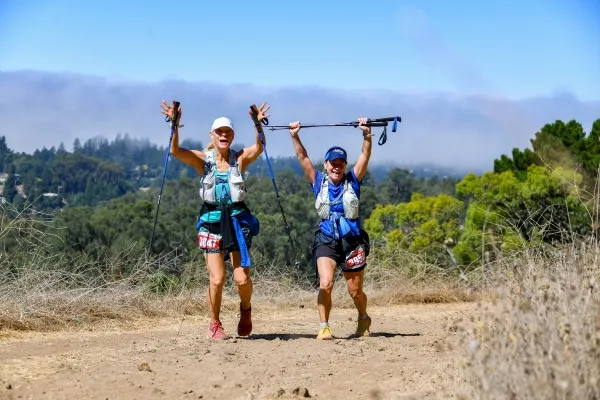 Two female runners with trekking poles raised in celebration on a sunny trail with rolling hills and trees in the background.