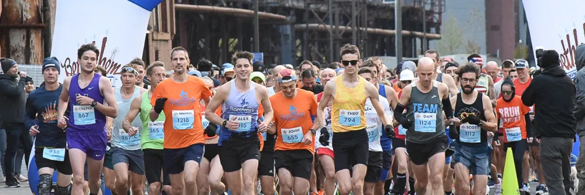 A group of runners in athletic wear, some with bib numbers, are starting a race. The lead runners are in focus, with a blurred background of industrial structures and banners.
