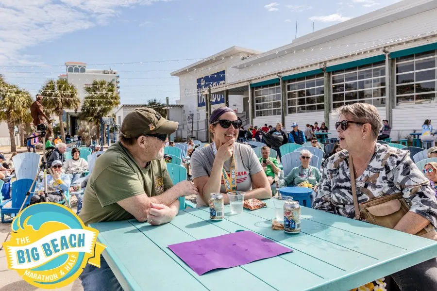People sitting at tables outside a building, some wearing race-related attire, with a beach and palm trees in the background.