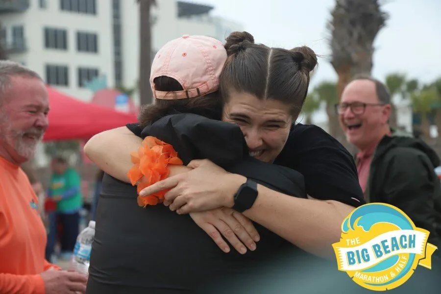 Two people hugging with smiles and laughter, with a race logo in the corner.