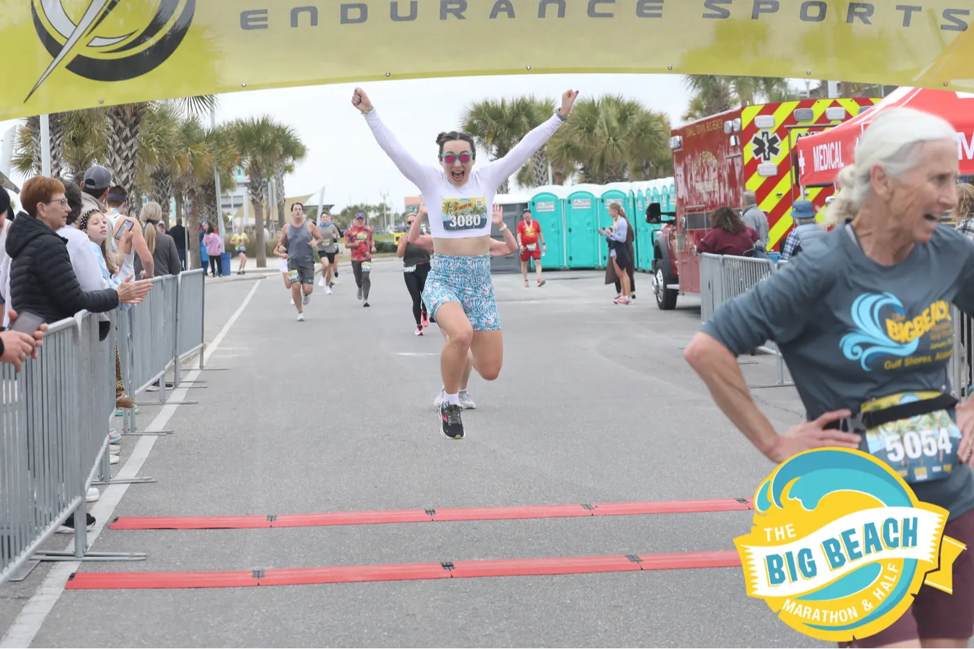 A female runner leaps across the finish line with her arms raised in celebration, while other runners and spectators watch.