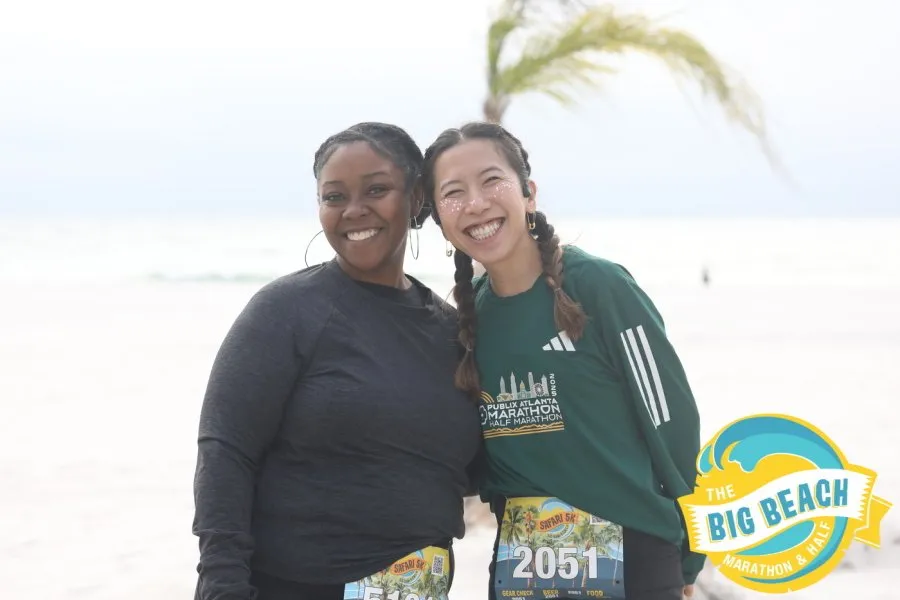 Two smiling women pose for a photo on a beach, wearing race bibs and race shirts.