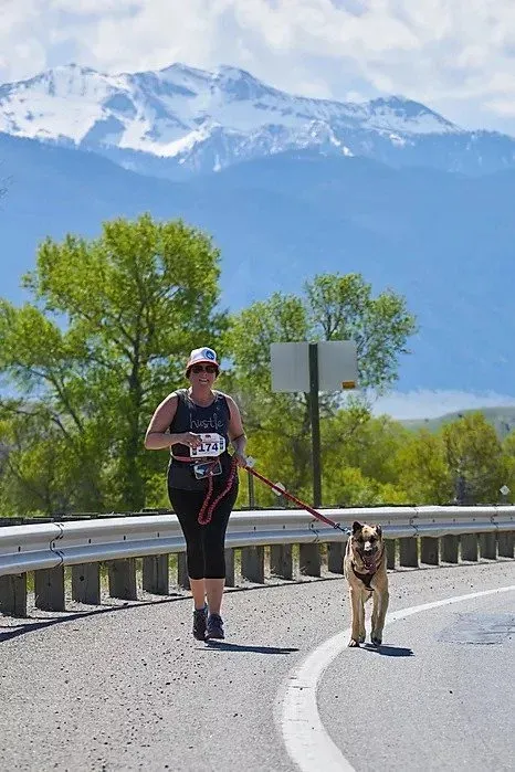 A woman runs with her dog on a road with snow-capped mountains in the background.