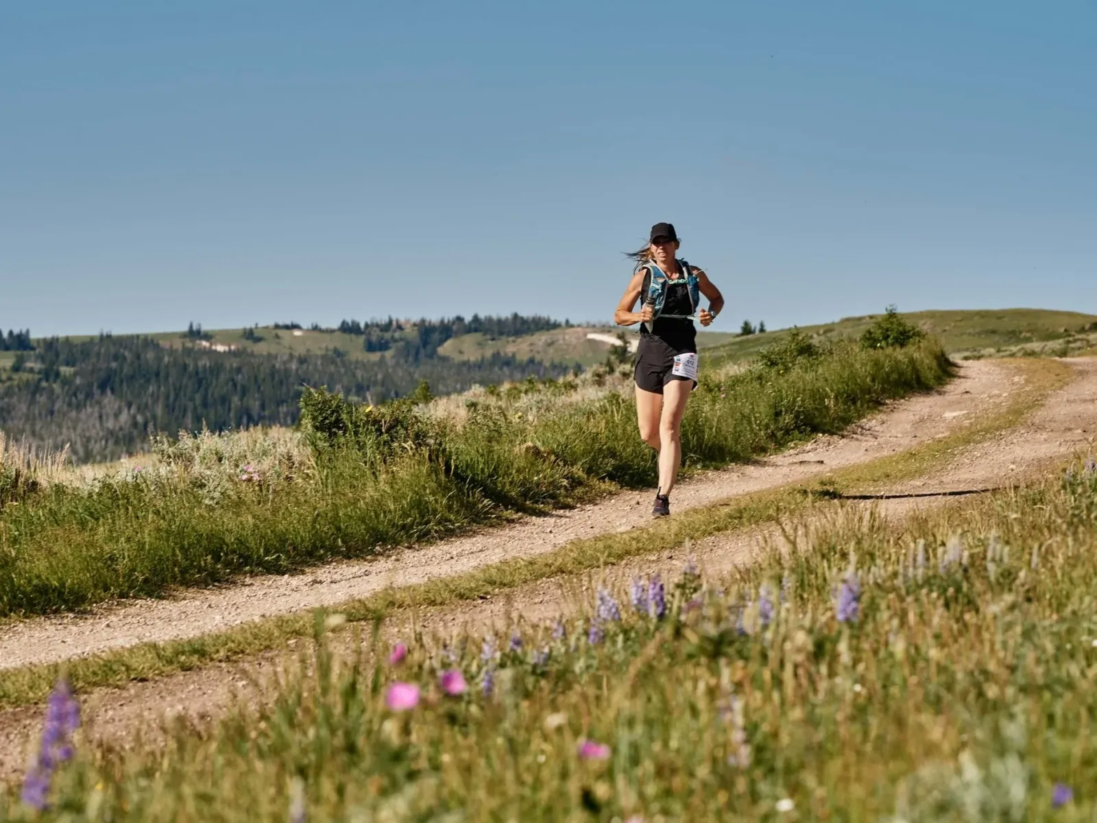 A female runner with a hydration pack runs on a dirt trail through a grassy, hilly landscape under a clear blue sky.