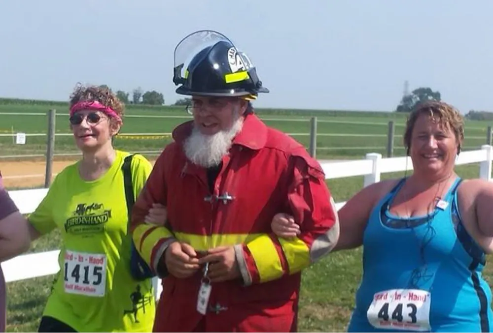 Three people, one dressed as a firefighter, walking together at a race event. The person on the left wears a green shirt with bib number 1415, and the person on the right wears a blue tank top with bib number 443.