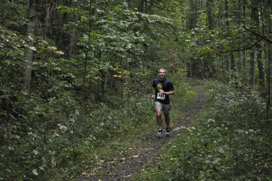 A male runner with bib number 484 smiles as he runs on a wooded trail.