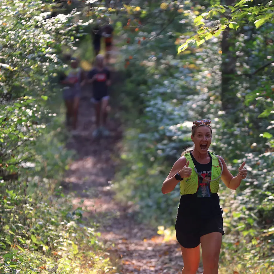 A woman with an excited expression and thumbs up runs on a wooded trail during a race.