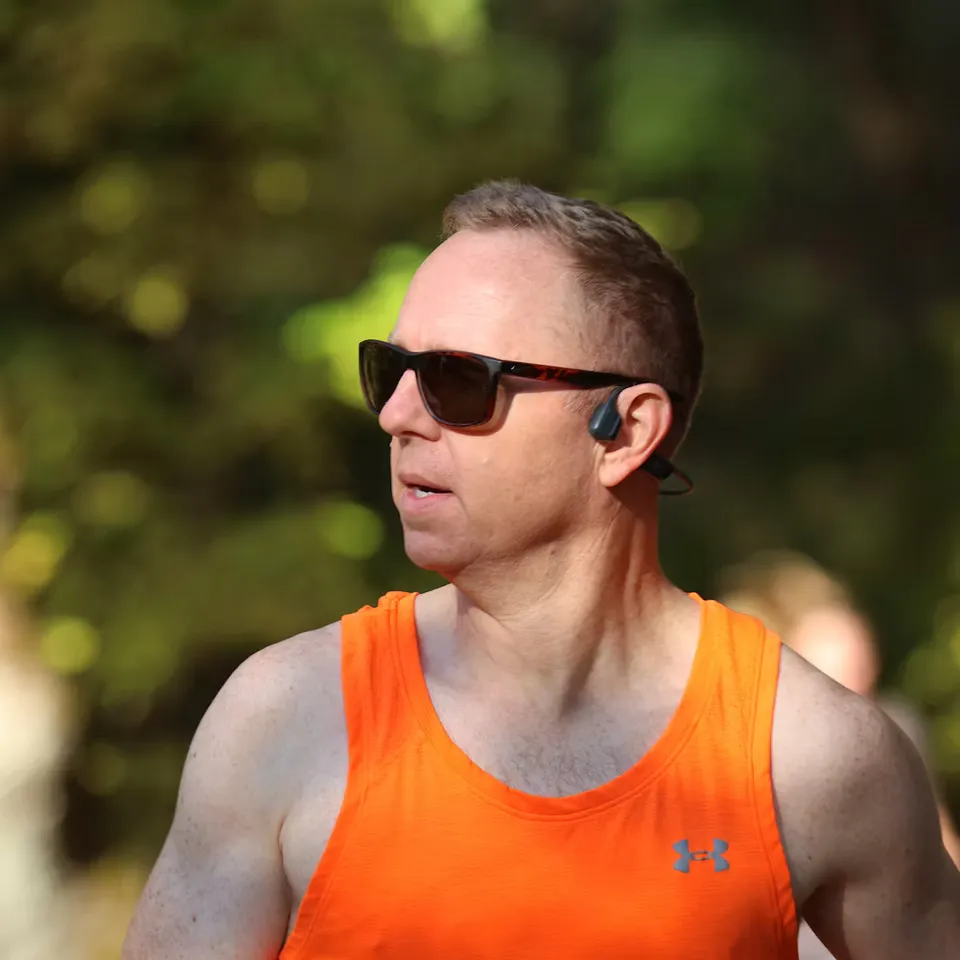 A male runner wearing sunglasses and an orange tank top, with a blurred background of trees.