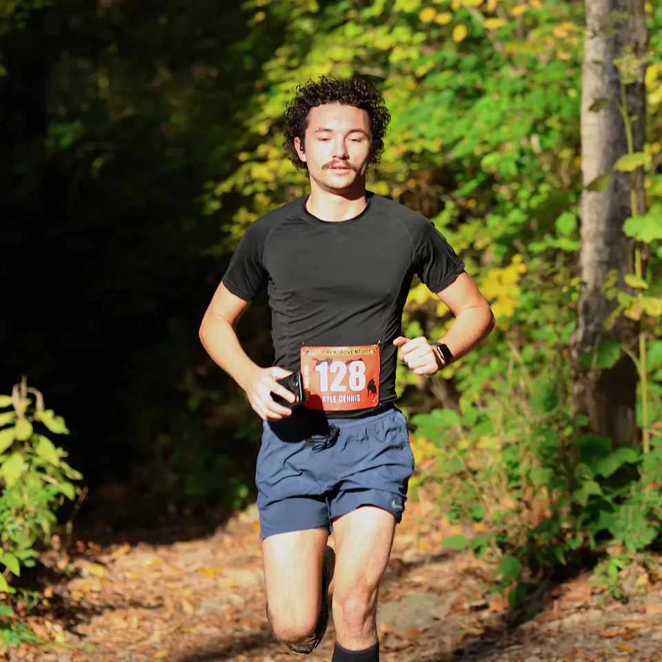 A male runner with curly hair and a mustache runs on a wooded trail, wearing a black shirt, blue shorts, and bib number 128.